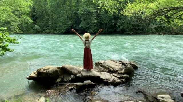 Young beautiful woman in long skirt and straw boater hat standing on stones near powerful rippling mountain river and raises her hands up. Summer travel, romantic vintage girl, old retro style clothes