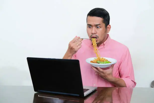 Adult Asian man looking to his laptop with shocked expression while eating noodle