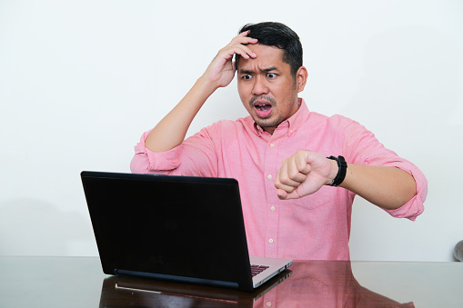 Adult Asian man looking to his laptop with worried expression while showing his watch