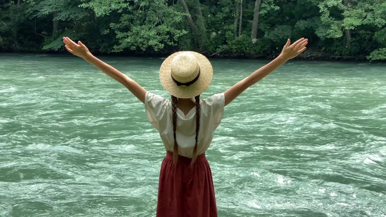 Young beautiful woman in long skirt and straw boater hat standing on stones near powerful rippling mountain river and raises her hands up. Summer travel, romantic vintage girl, old retro style clothes