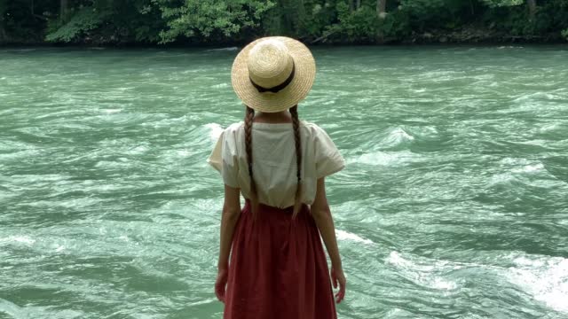Young beautiful woman in long skirt and straw boater hat standing on stones near powerful rippling mountain river. Summer travel concept, romantic vintage girl, old retro style clothes