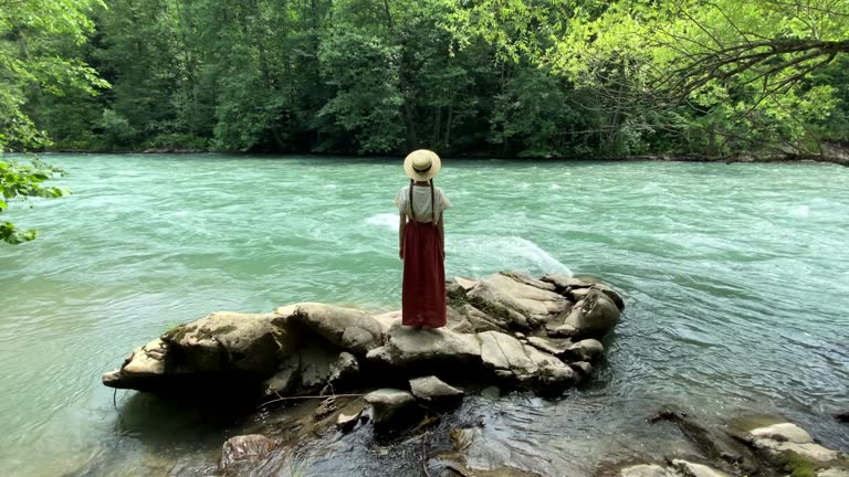 Young beautiful woman in long skirt and straw boater hat standing on stones near powerful rippling mountain river. Summer travel concept, romantic vintage girl, old retro style clothes