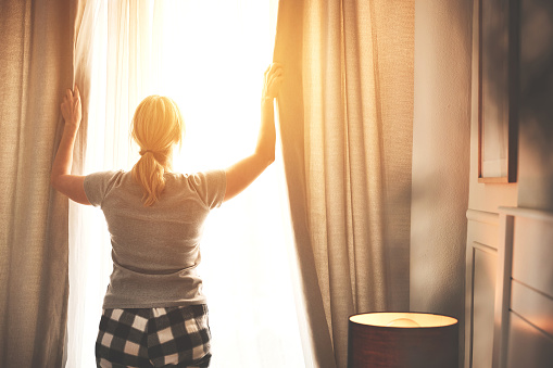 A woman opening the curtains on a bright morning at sunrise inside her bedroom. A female opens drapes to let the sunlight into her room. Lady wearing pajamas or PJs awake and ready for the day