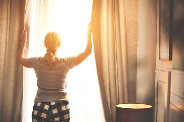 вид сзади женщины, открывающей шторы ярким летним утром на рассвете в своей спальне. самка открывает шторы, чтобы пропускать солнечный свет - waking up window women morning стоковые фото и изображения