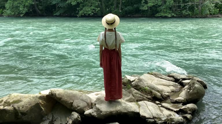 Young beautiful woman in long skirt and straw boater hat standing on stones near powerful rippling mountain river. Summer travel concept, romantic vintage girl, old retro style clothes