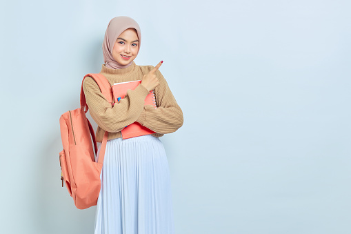 Smiling young Asian Muslim woman student in brown sweater with backpack, pointing fingers aside at copy space isolated on white background. back to school concept