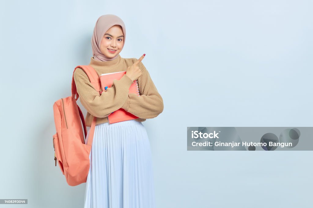 Smiling young Asian Muslim woman student in brown sweater with backpack, pointing fingers aside at copy space isolated on white background. back to school concept Student Stock Photo