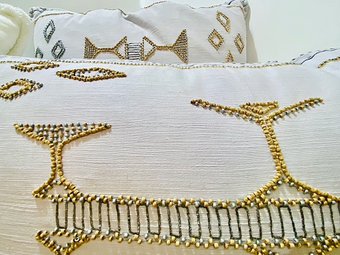 Horizontal close up of gold and silver bead design embroidered on white throw lounge cushions in interior room