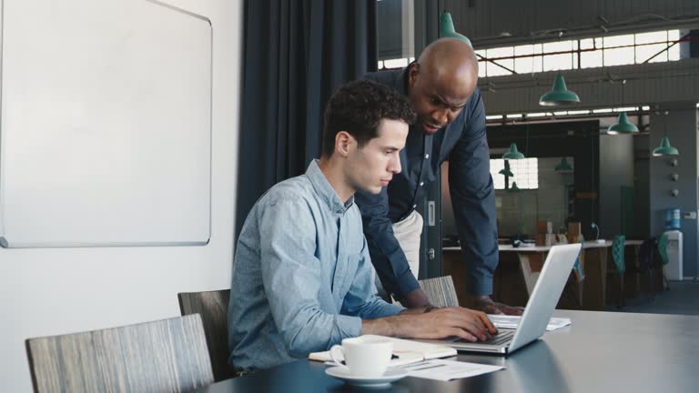 Two businesspeople discussing something on a laptop. Intern showing computer presentation to skilled team leader. Diverse colleagues working together using app. Having a meeting and planning online