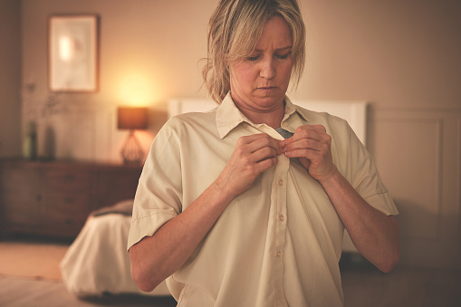 Mature woman getting dressed and buttoning her shirt in a bedroom in the morning. Female putting on fresh clothing while preparing for the day. Freshening up and getting ready to leave the house