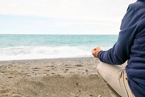 Rear view of man in casual clothes sitting in lotus pose on pebble winter beach and meditating on cloudy day. Healthy lifestyle. Yoga concept
