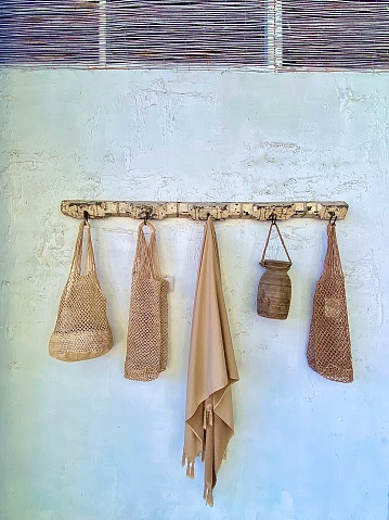 Horizontal close up of domestic hallway white rustic wall with wood hook rail rack holding tan beach towel and handmade woven and wood carry bags