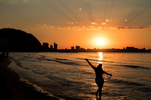 Girl posing with her arms up in a sunrise at the postiguet beach in Alicante, with buildings against the light.