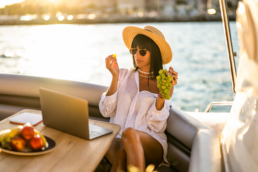 Female business person with sunglasses and a hat in a white shirt seen holding and eating grapes while having a video call on her laptop during a leisure trip on a yacht where she is also working at the same time.