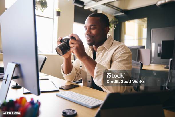 Young Black Professional Photographer Sitting At Desk Working On Computer Holding Camera Editing Pictures Stock Photo - Download Image Now