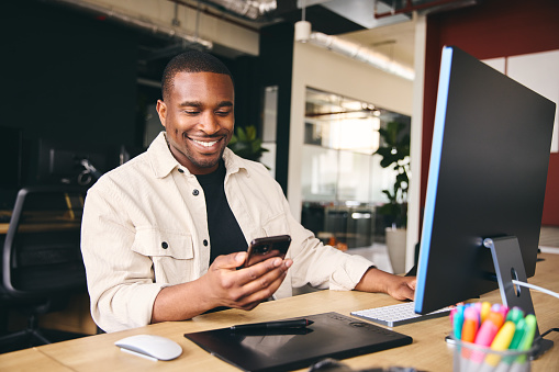 Young Black Male Advertising Marketing Or Design Creative In Modern Office Sitting At Desk Using Mobile Phone