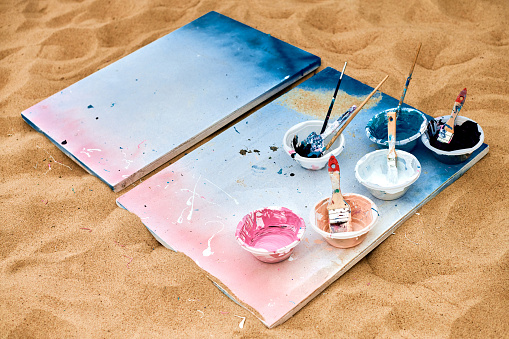 Two painted canvas in blue and pink colors with palettes of paints lying on sandy beach, artist's palette at outdoor art festival. White drawing canvas with paint in bowls, art performance concept