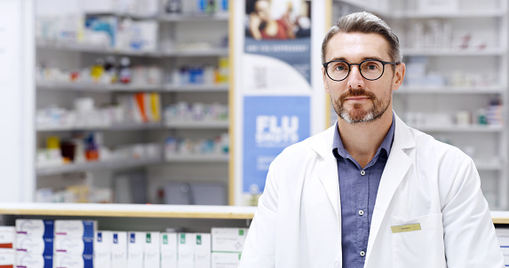 Portrait of confident male pharmacist in a drugstore. Caucasian healthcare professional in a pharmacy looking at the camera. Portrait of a doctor standing in a clinic alone wearing a white coat