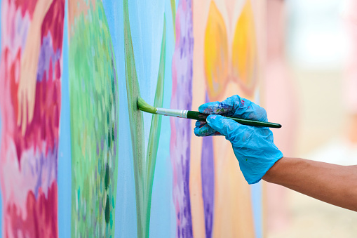 Artist's hand with paintbrush painting colorful picture on canvas at outdoor art festival. Woman painter in blue gloves draws surreal picture with paintbrush, beautiful art performance