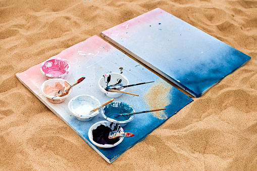 Two painted canvas in blue and pink colors with palettes of paints lying on sandy beach, artist's palette at outdoor art festival. White drawing canvas with paint in bowls, art performance concept
