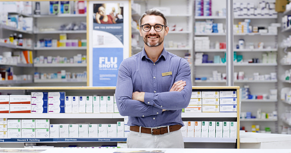 Portrait of a happy pharmacist standing with arms crossed in a pharmacy. Mature expert healthcare worker with a proud smile while behind a counter with a variety of medical supplies in the background