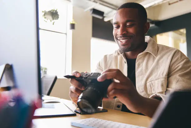 Photo of Young Black Professional Photographer Sitting At Desk Working On Computer Holding Camera Editing Pictures