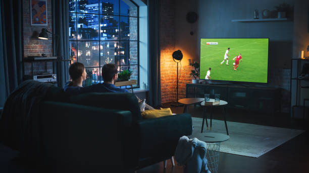 Couple of Soccer Fans Relax on a Couch, Watch a Sports Match at Home in Stylish Loft Apartment. Relaxed young Man and Woman Cheer for Their Favorite Football Club and Enjoying the Evening. Couple of Soccer Fans Relax on a Couch, Watch a Sports Match at Home in Stylish Loft Apartment. Relaxed young Man and Woman Cheer for Their Favorite Football Club and Enjoying the Evening. tv game stock pictures, royalty-free photos & images