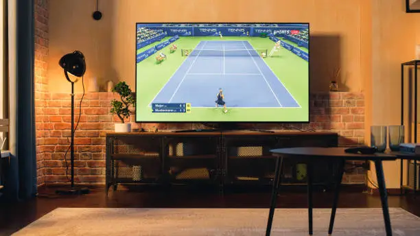 Photo of Stylish Loft Apartment Interior with Tennis Game Playing on Flat Screen Television. Empty Living Room at Home With Broadcast of Two Female Professional Tennis Players Competing. Sunset Shot.