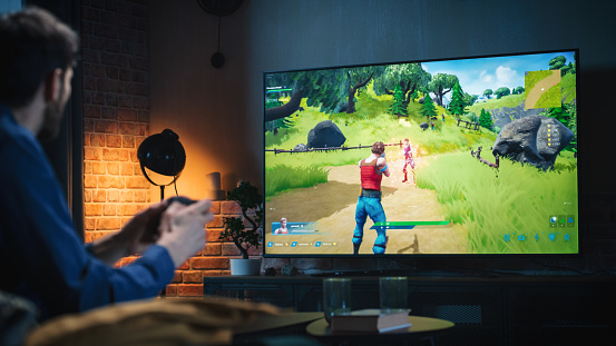 Young Man Spending Time at Home, Sitting on a Couch in Stylish Loft Apartment and Playing Arcade Shooter Video Game on Console. Male Using Controller to Play MMO Battle Royale Style Game Online.