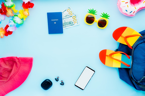 Top view summer vacation concept with all necessary travel stuff. Backpack, bucket hat, funny sunglasses, flip flops, biometric passport, cash, phone with blank screen, ear airpods. Copy space.