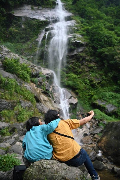 Mother and daughter taking a selfie with mobile phone while sitting on a rock near Changey waterfall in Sikkim