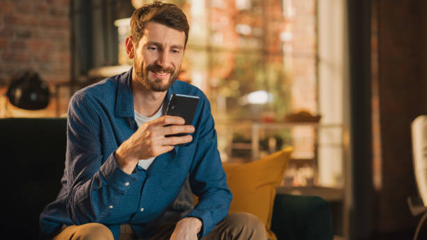 Portrait of a Handsome Man Sitting on Couch, Using Smartphone in Stylish Loft Apartment or Cafe. Creative Male Smiling, Checking Social Media and Typing Message. Urban City View from Big Window. stock photo