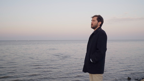 young man with overcoat looks at the horizon at sunset next to the river