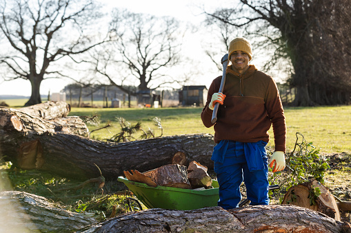 A front-view portrait shot of a young man working on a farm, he is smiling and looking at the camera while holding an axe over his shoulder, he has been collecting wood in a wheelbarrow after trees have been damaged during a storm in Northumberland.