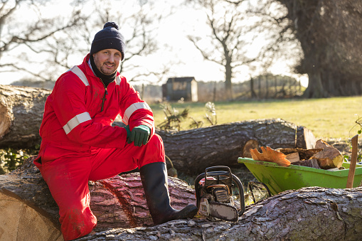 A front-view portrait shot of a mid-adult man working on a farm, he is smiling and looking at the camera while sitting on a wooden log, he has been collecting wood in a wheelbarrow after trees have been damaged during a storm in Northumberland.