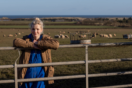 A front-view three-quarter-length shot of a young female farmer on a field with a flock of sheep  behind her at a farm in Northumberland.
