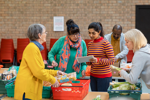 Group of volunteers organising food donations onto tables at a food bank in the North East of England. They are working together looking at a digital tablet while setting up sections of the room in a church.