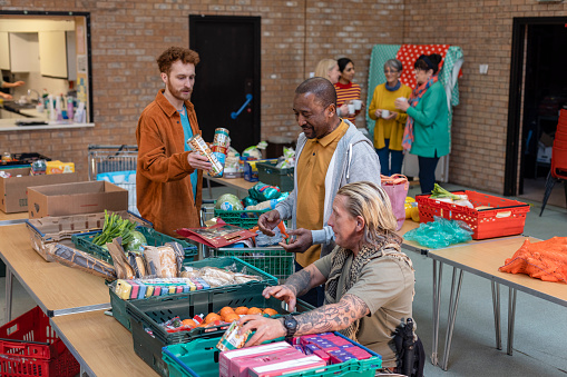 Male group of volunteers organising food donations onto tables at a food bank in the North East of England. They are working together, setting up sections of the room in a church. A group of women are drinking hot drinks out of focus in the background.