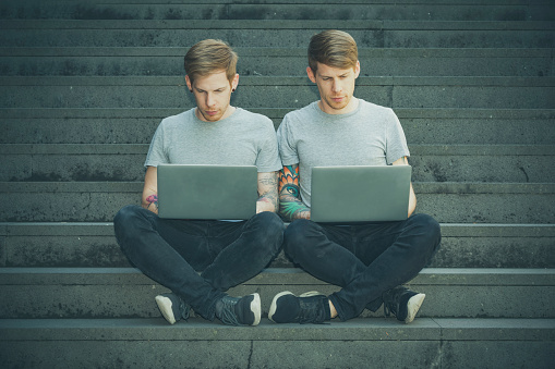 twin brothers sitting next to each other working on laptops in urban surrounding.