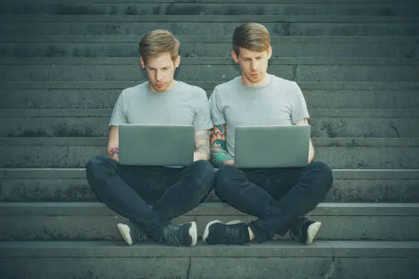 twin brothers sitting next to each other working on laptops in urban surrounding.