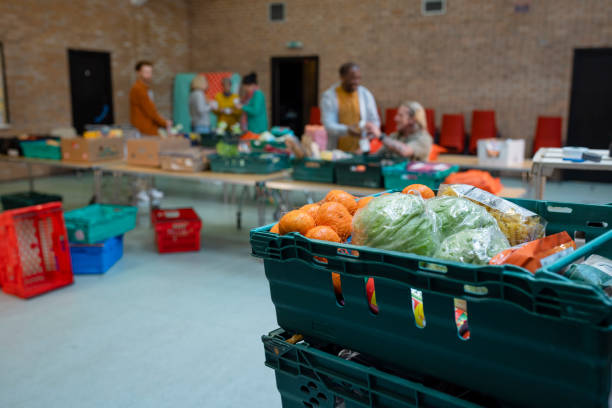 Donations at a Food Bank Charity Selective focus image showing fresh fruit and vegetable food donations in crates at a food bank in the North East of England. There are out of focus volunteers working together in the background, setting up the event. food bank stock pictures, royalty-free photos & images