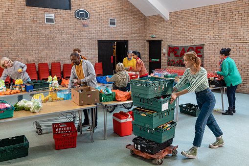 Group of volunteers organising food donations onto tables at a food bank in the North East of England. They are working together, setting up sections of the room in a church. One woman is pushing stacked grates.