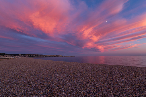 Pink sky as the sun set at Chesil beach with bank of shingles and flat calm sea reflecting the pink glowing clouds along the Jurassic coast of Dorset.