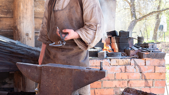 A medieval blacksmith in a traditional clothes with a leather apron forges a horseshoe on an anvil. The blacksmith stands at the forge furnace and heats the metal. Medieval professions concept.