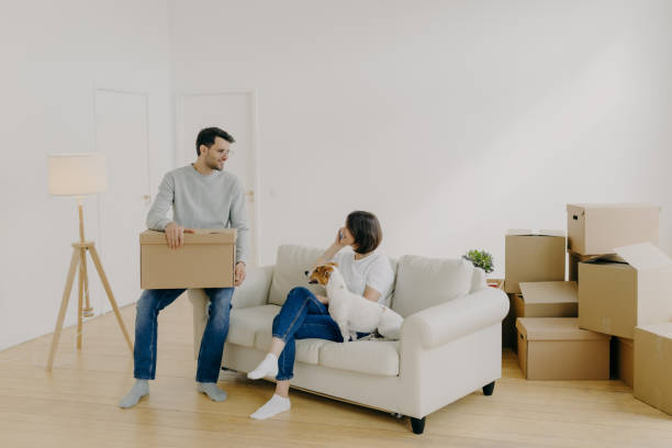 istock Positive woman and man pose in empty spacious room during relocation day, husband carries cardboard boxes with belongings, wife has telephone conversation, sits on comfortable sofa with dog. 1408438761