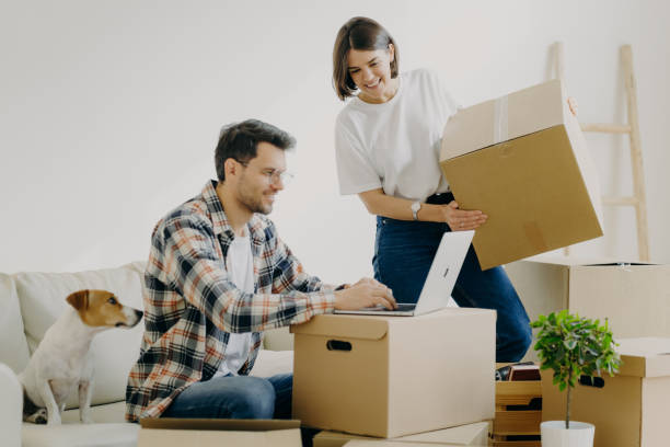 istock Image of happy young man keyboards something on laptop computer, has glad expression, sits on comfortable sofa with pet, surrounded with cardboard boxes, make online shopping, buy new furniture 1408438767