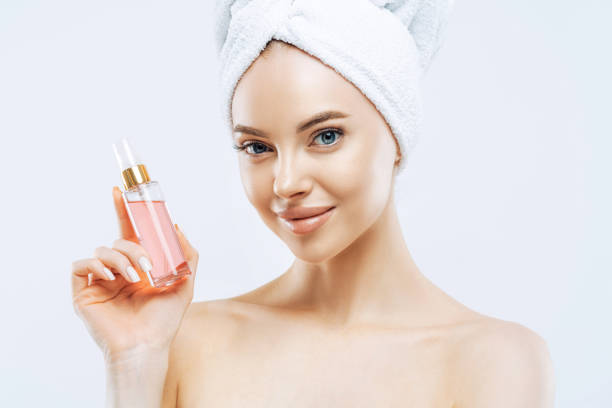 attraente giovane donna applica parfum, gode di un profumo piacevole, sta con le spalle nude, ha trucco naturale, pelle sana, asciugamano avvolto sulla testa dopo aver fatto la doccia. ottimo aroma, prova questo. - abbigliamento intimo foto e immagini stock