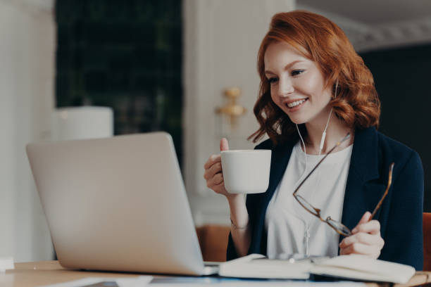 istock Intelligent female student has online course, concentrated in screen of laptop computer, learns foreign languages distantly, drinks tea or coffee, poses at desktop. Woman worker has video conference 1408441906