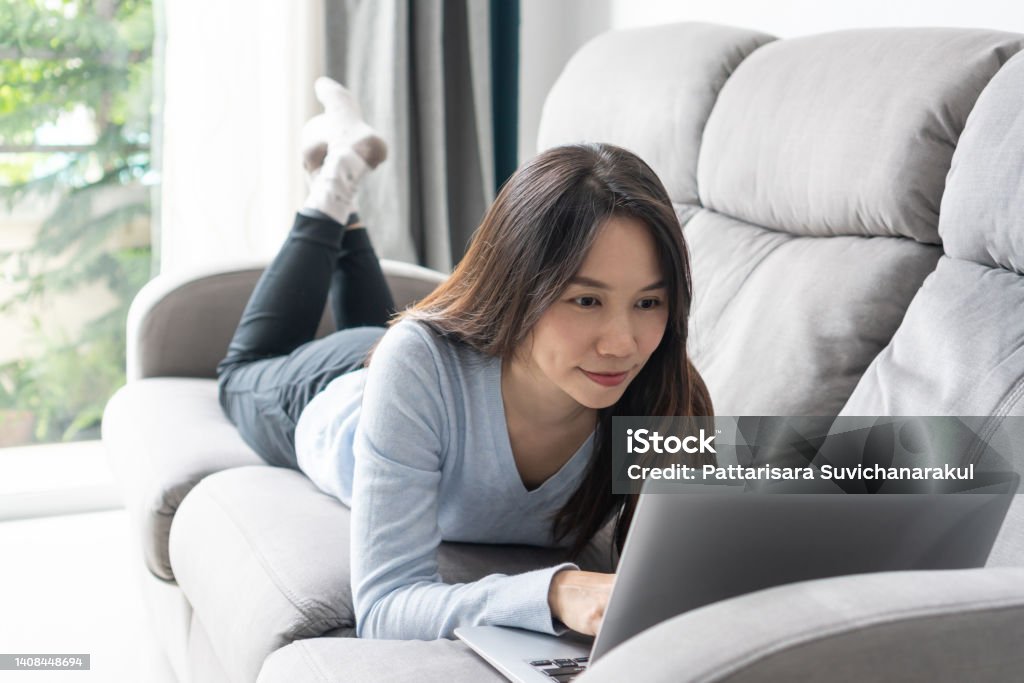 Happy woman lying down on sofa using laptop communicating online at home. Smiling girl working on computer, enjoying internet shopping or chatting in social networks. Technology and lifestyle concept. Laptop Stock Photo