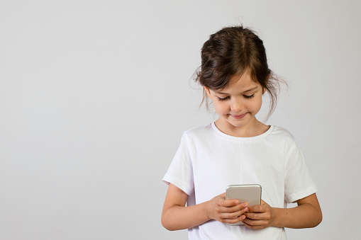 6 years old girl standing against white background while looking to mobile phone.